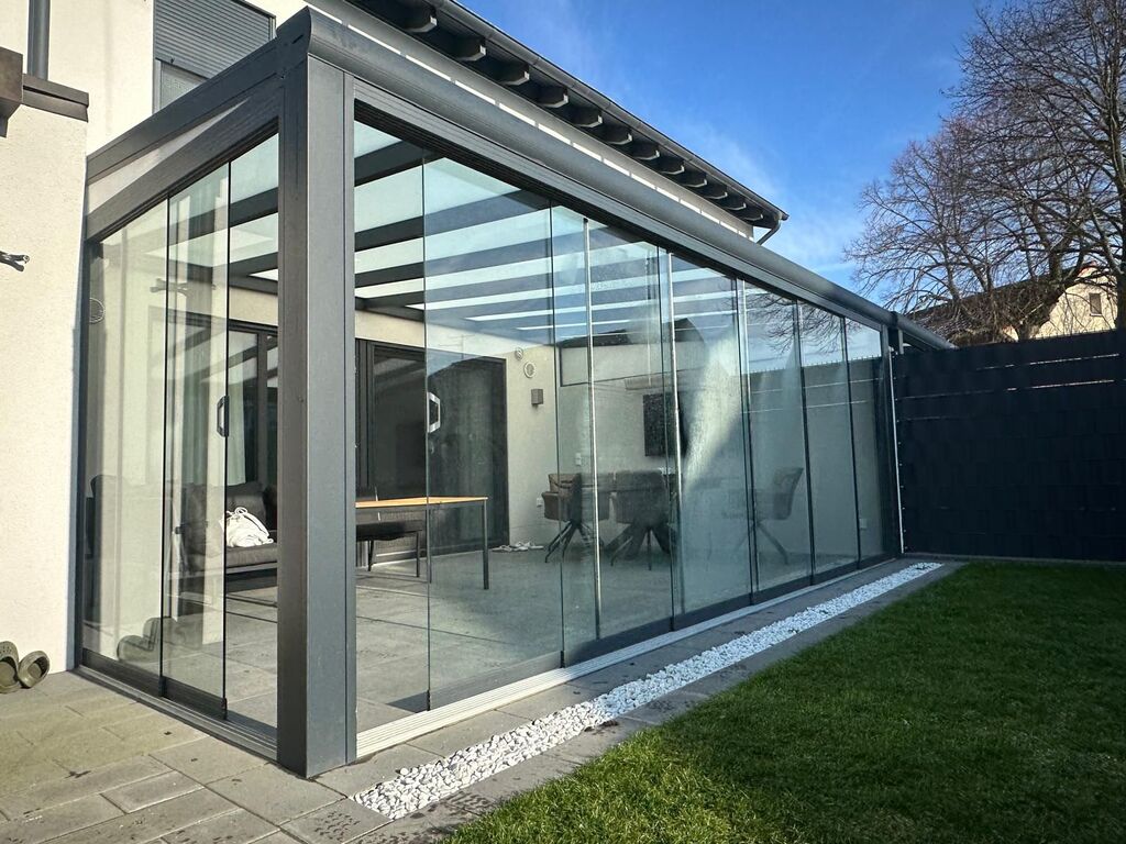 Moderner Wintergarten mit Glaswänden, grauem Rahmen, an Haus neben Rasen und kieselsteinbedecktem Rand.