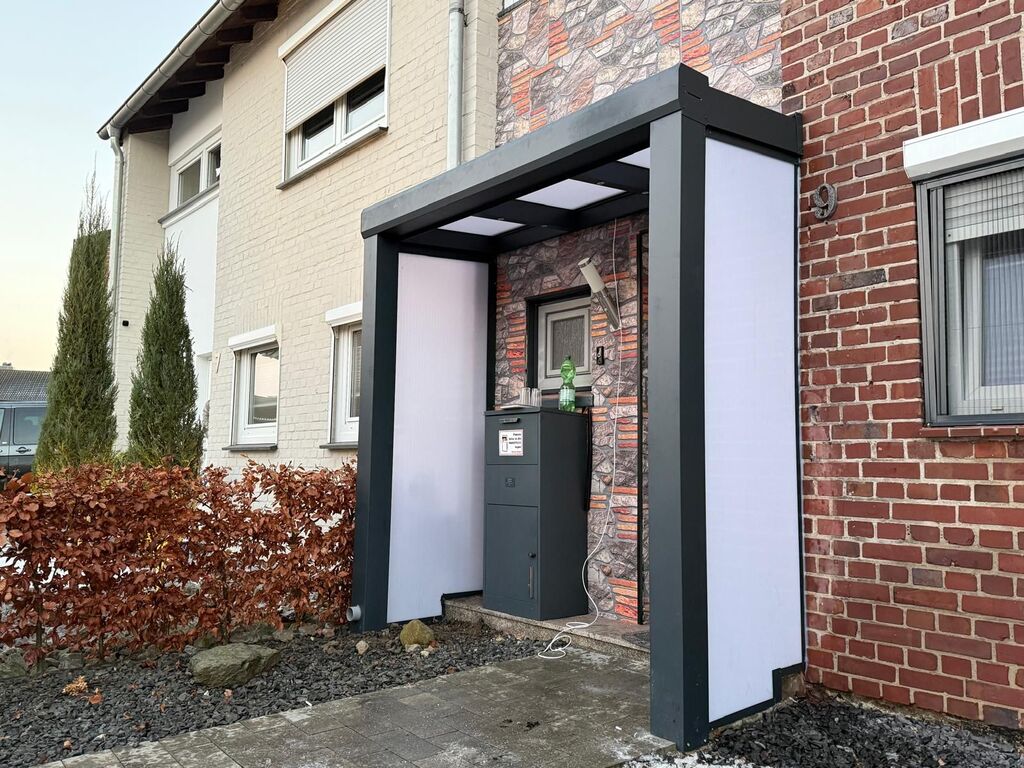 Ein moderner Eingang mit Vordach, weiß und anthrazit, mit Ziegelwand und dekorativem Strauch.