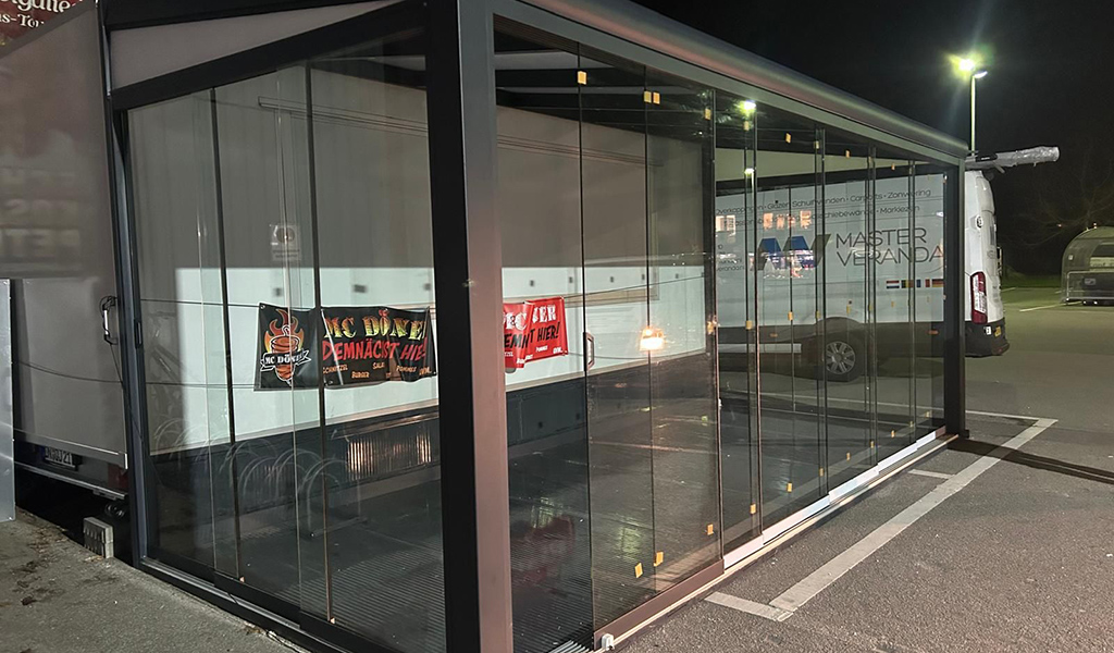Ein leerer verglaster Pavillon mit Werbebannern vor einem Lieferwagen in einem dunklen Parkplatz.