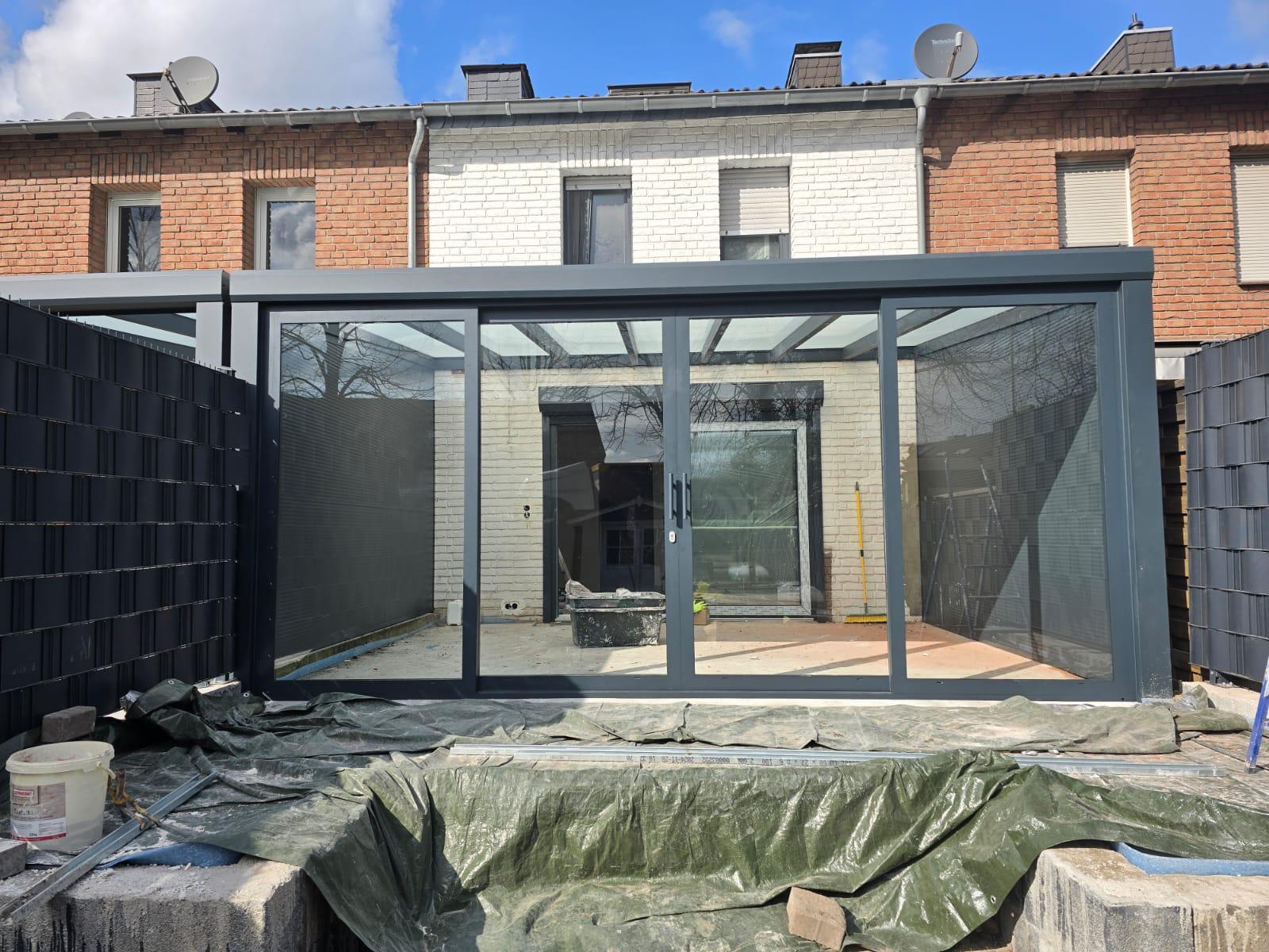 Moderner Wintergarten-Anbau an einem Backsteinhaus mit großen Glasfenstern und grauem Rahmen.