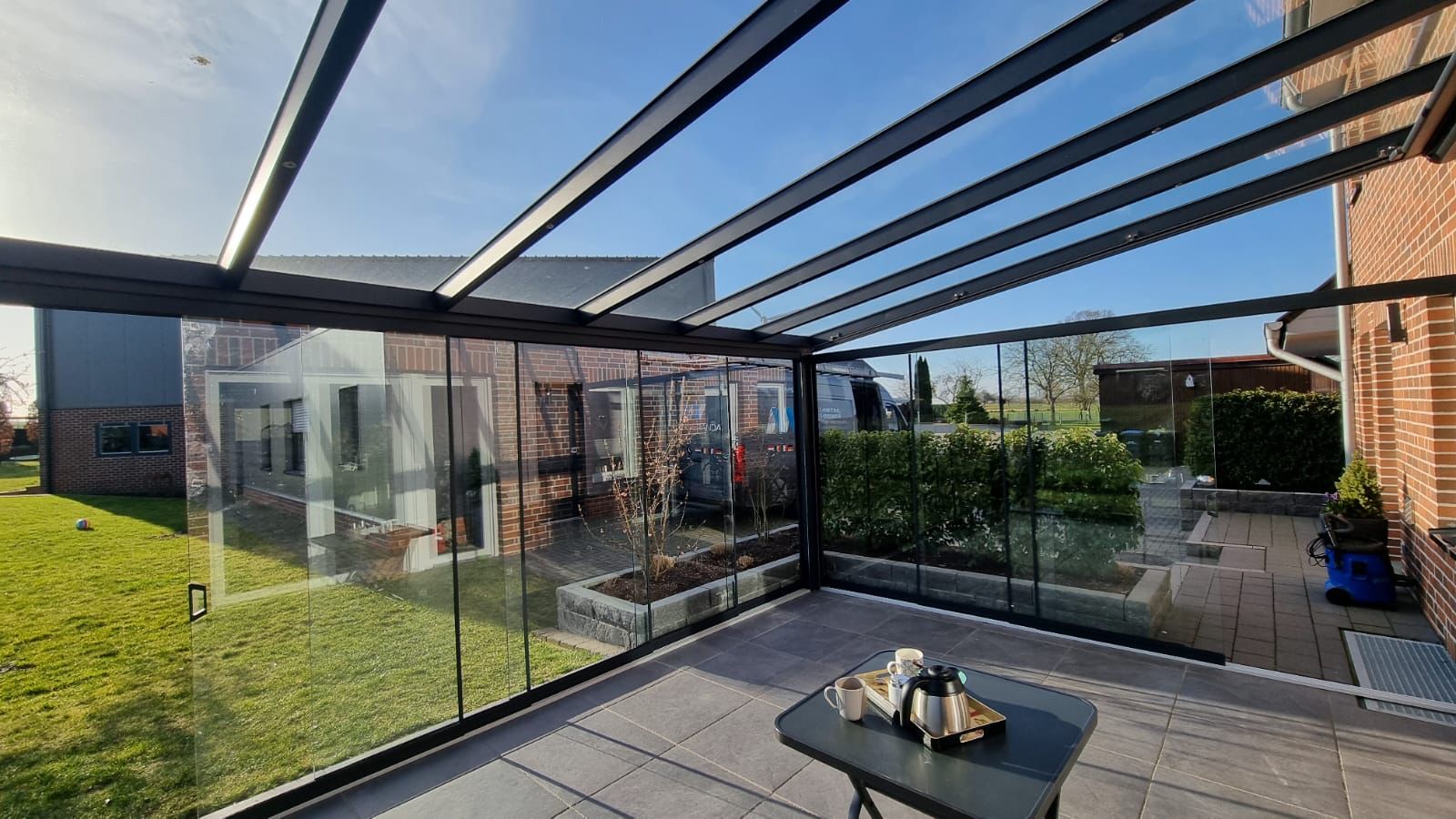 Moderne Glasveranda mit Tisch, Teekanne; sonniger Tag, Gebäude und Garten im Hintergrund.