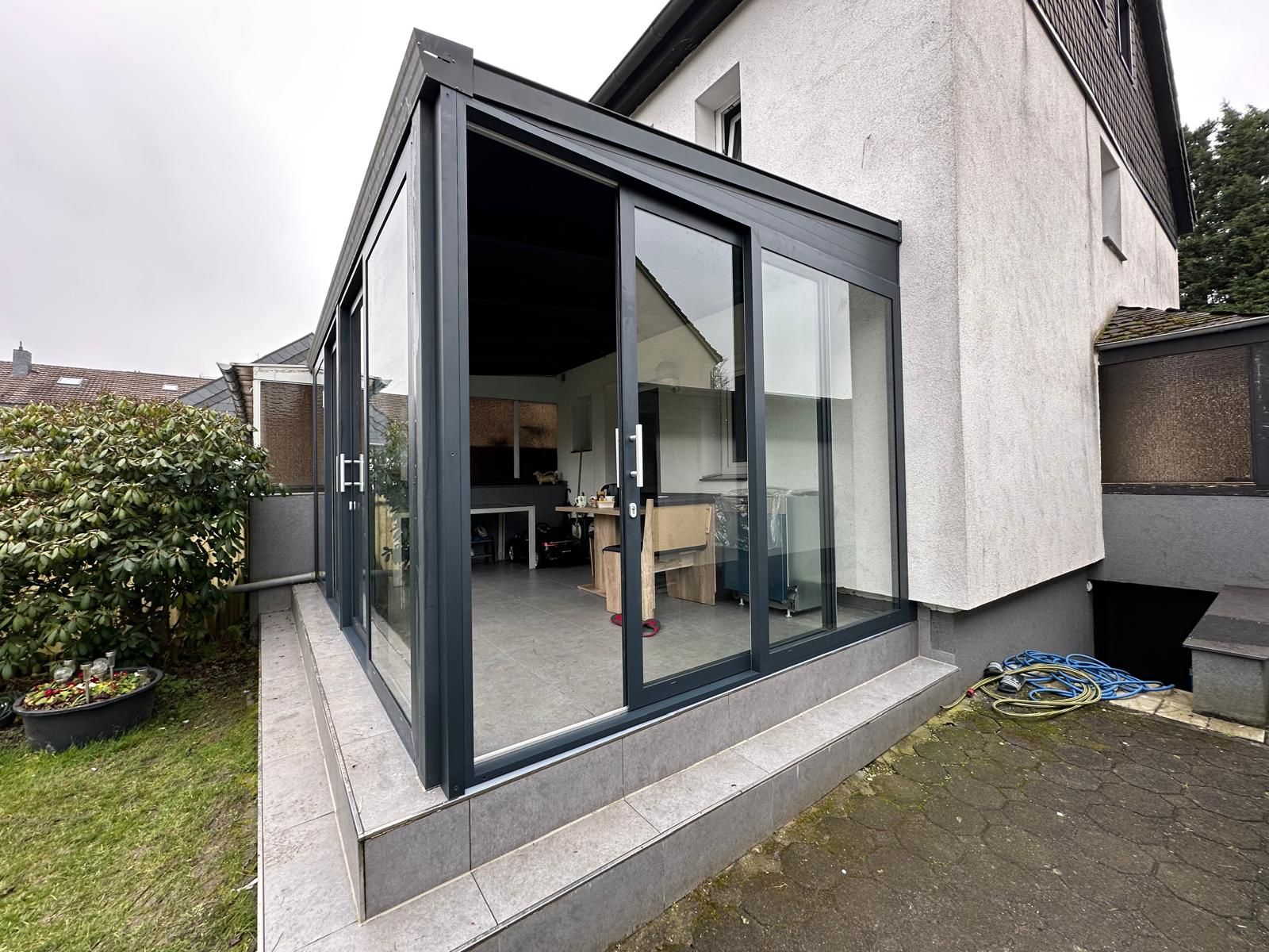 Ein modernes Glas-Wintergarten-Anbau mit Aluminiumrahmen an einem grauweiß gestrichenen Haus.