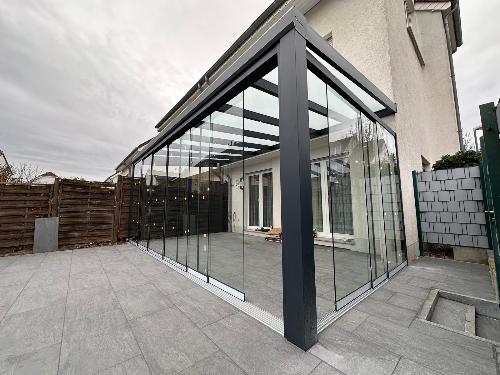 Veranda mit Glaswänden, graue Fliesen, modernes Design, bewölkter Himmel im Hintergrund.