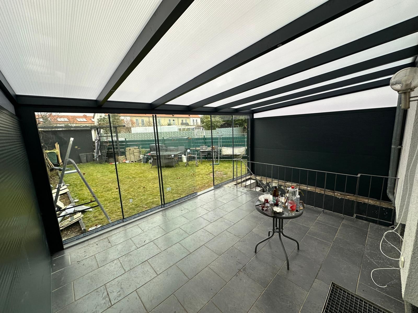 Überdachte Terrasse mit Glaswänden, Blick auf Garten, Tisch mit Flaschen, Schaukeln im Hintergrund.