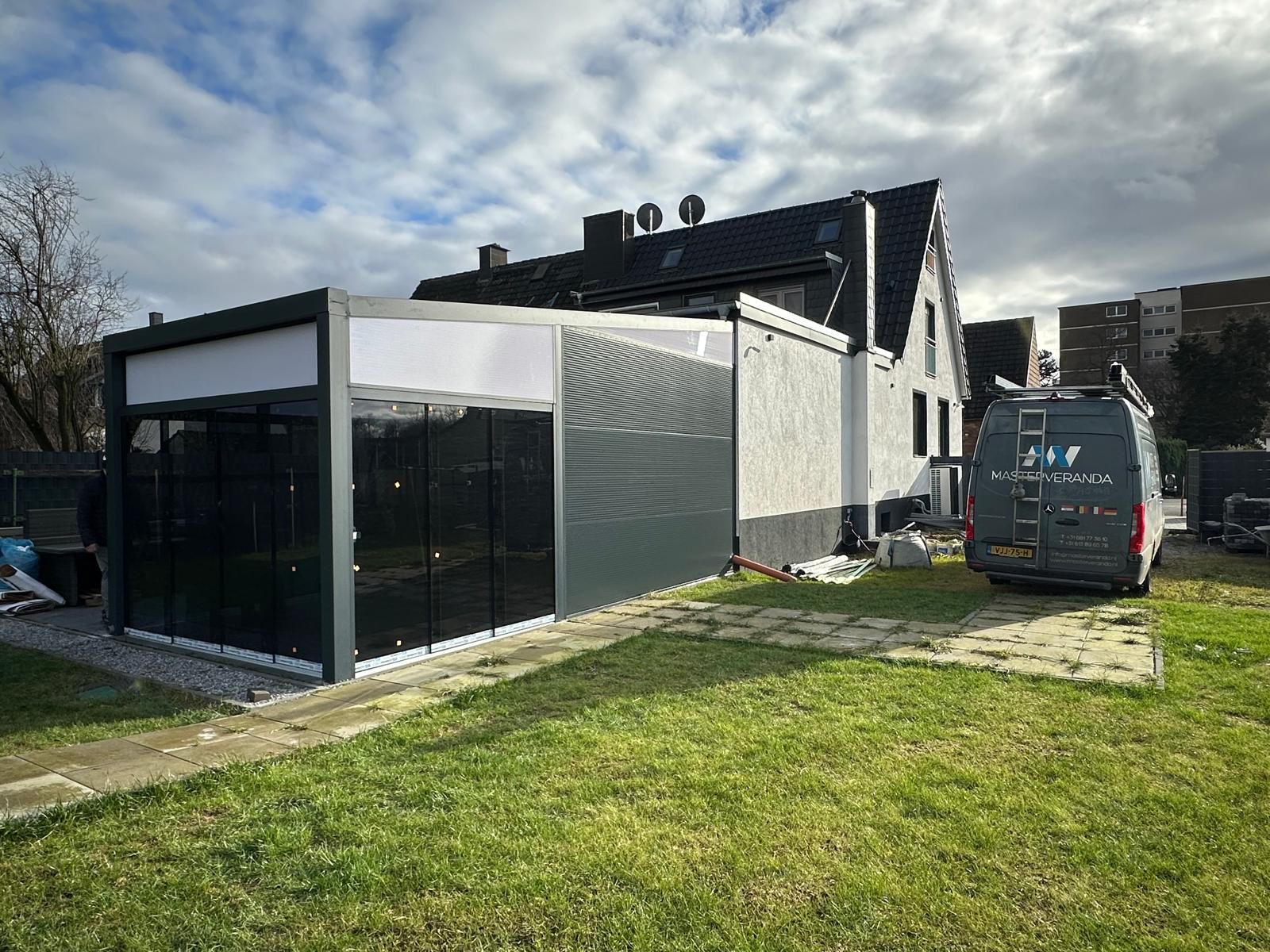 Moderner Anbau mit Glasfront, grauem Haus, daneben weißer Firmenwagen auf grünem Rasen.