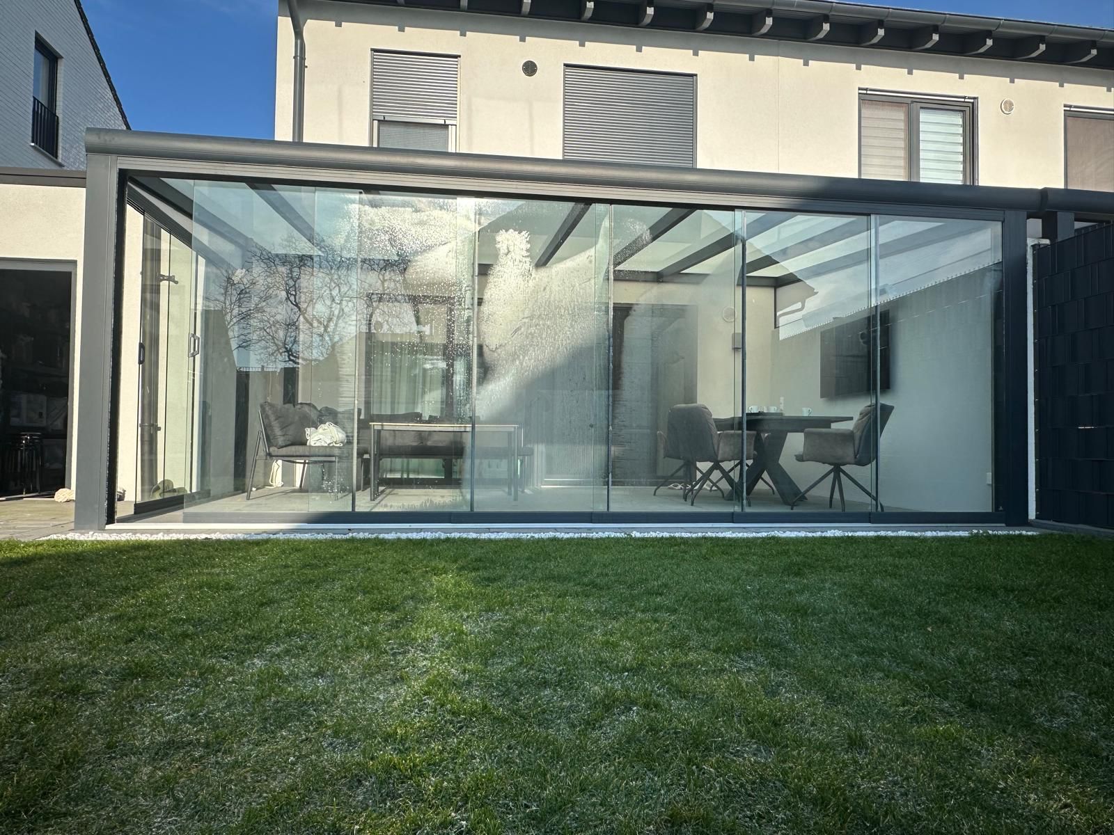 Moderne verglaste Veranda mit Esstisch, graue Stühle; dahinter zweistöckiges Haus, blauer Himmel.
