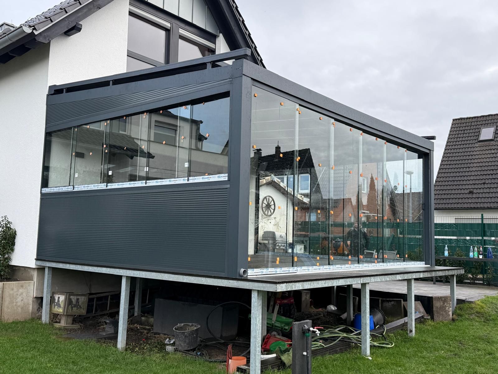 Modernes graues Terrassenzimmer mit Glaswänden; darunter Stauraum, grüne Wiese vorne.