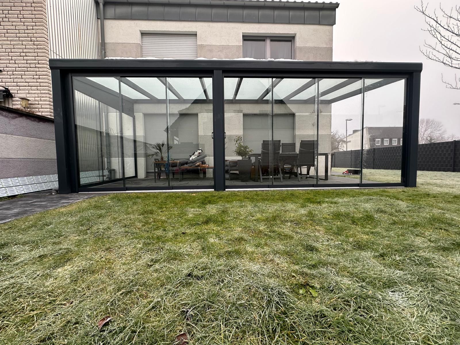 Geschlossener Wintergarten mit Tisch und Stühlen, aus Glas und Metall, auf einem frostigen Rasen.