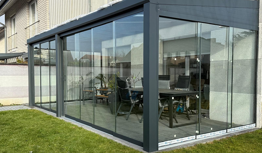 Moderne, verglaste Veranda mit grauem Rahmen und Blick auf Tisch und Stühle im Inneren.